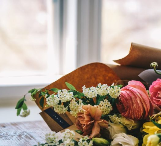 Blommor i olika färger och sorter ligger på ett bord framför ett fönster, inslagna i brunt papper.